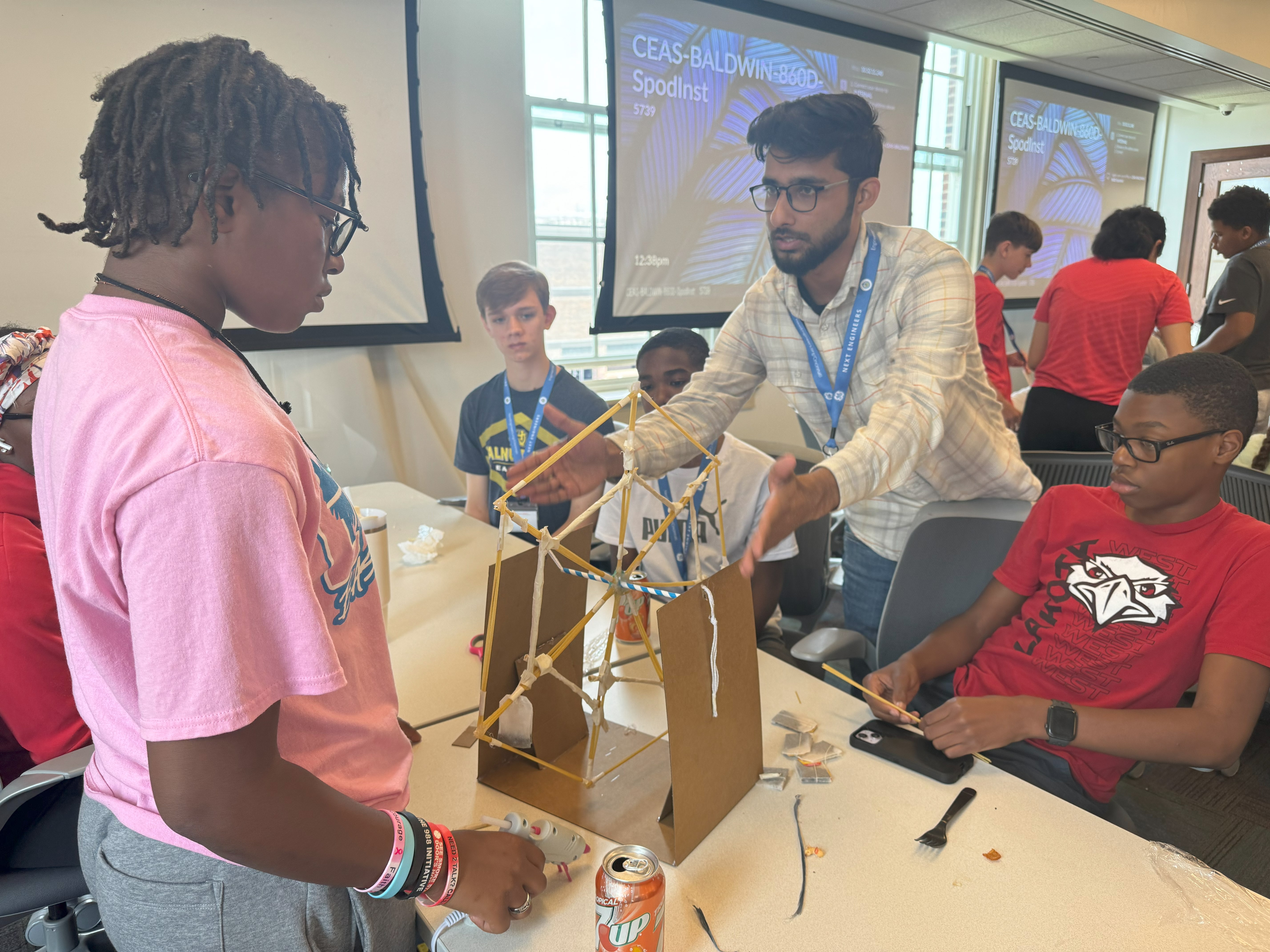 Counselor Aseem, in a plaid shirt, holds his hands over a Ferris wheel of pasta to assist students in their construction. The students look at the wheel as he talks. An African American girl stands across from him in a pink shirt. 