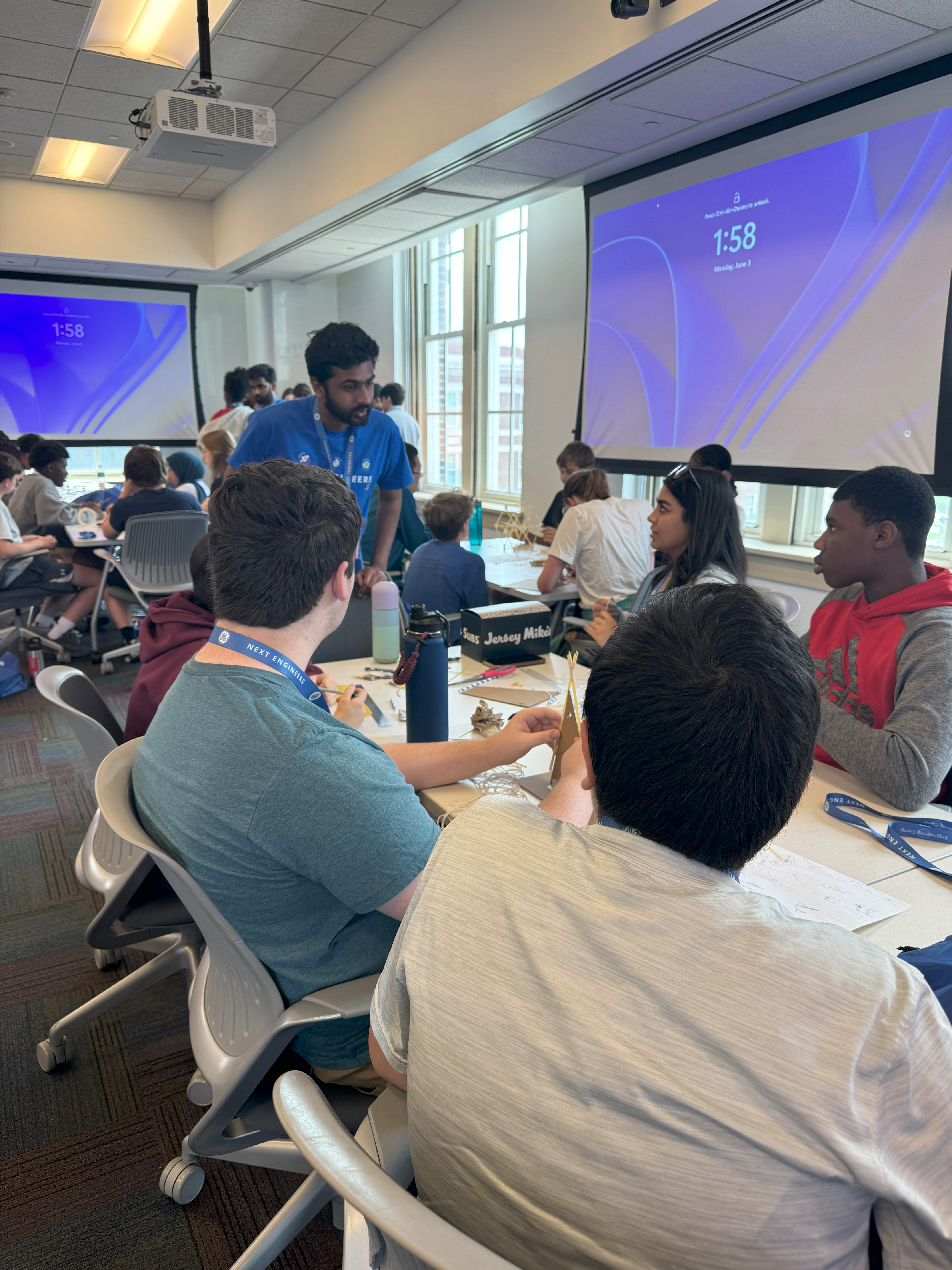 Counselor Sree talks with students about their project. He stands at the end of the table in blue "GE Next Engineers" attire while the students turn to him and look