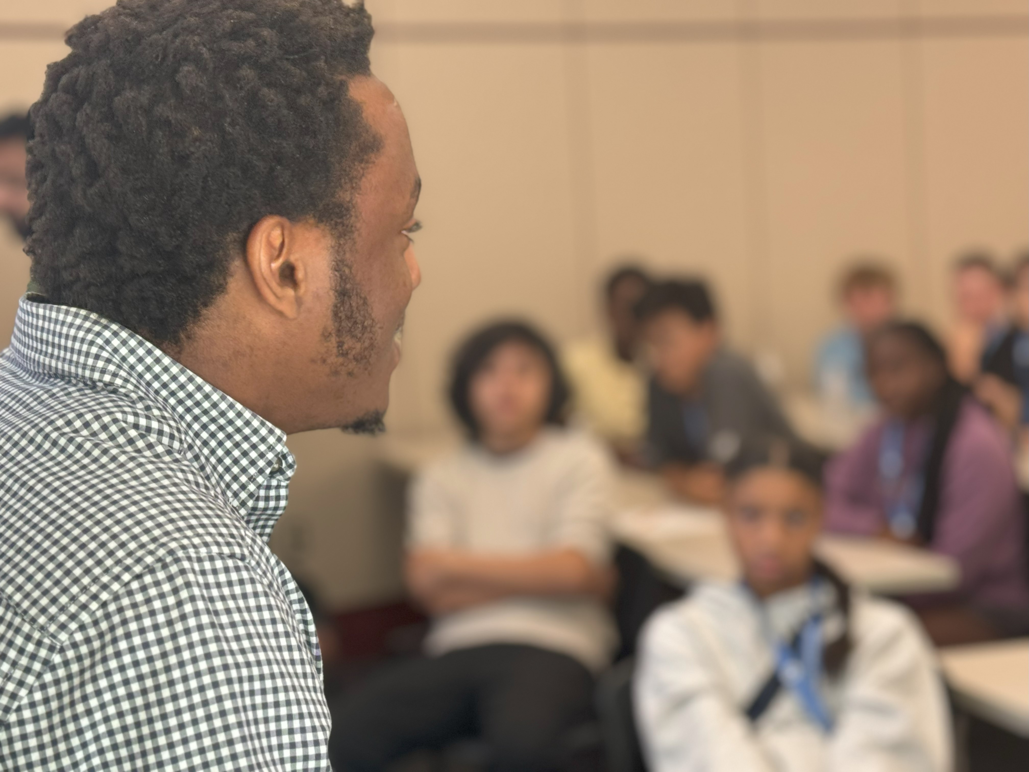 Camp Facilitator Okechukwu addresses the class in a plaid shirt. The students, just out of focus, look at him as he speaks. 